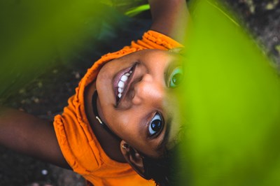 An overhead, close-up shot captures a young child smiling brightly while peeking up through vibrant green foliage. The child has large, dark eyes, a joyful grin showing white teeth, and wears a textured orange t-shirt. Large, out-of-focus bright green leaves flank the right and top edges of the frame, partially obscuring the view and creating a playful, natural border. 