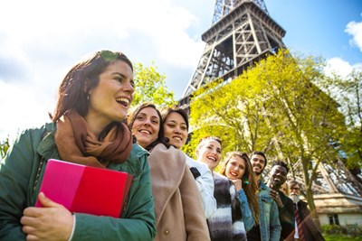A low-angle, wide shot captures a diverse group of students smiling and laughing as they stand lined up in a row outdoors, with the Eiffel Tower towering in the background. In the foreground, a woman wearing a green jacket and a brown scarf smiles while clutching a bright pink and red notebook. The other students behind her follow in a diagonal line, framed by green trees under a bright blue sky. 